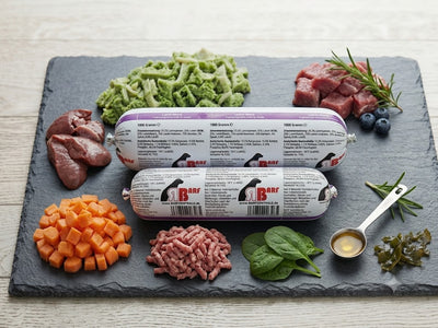 "Professionelles Produktbild des 'Hausmarke Lamm Menü' Hundefutters von BARF-Zentrale. Zwei gestapelte BARF-Rollen (1000g und 500g) liegen auf einer dunklen Schieferplatte. Sie sind umgeben von frischem Lammfleisch, gewürfelten Karotten, Muskelfleisch und einem Löffel Lachsöl."