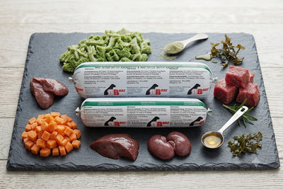 Professionelles Produktbild des 'Hausmarke Pansen Menü' Hundefutters von BARF-Zentrale. Zwei gestapelte BARF-Rollen (1000g und 500g) liegen auf einer dunklen Schieferplatte. Sie sind umgeben von frischem grünem Pansen, gewürfelten Karotten, rohem Muskelfleisch und einem Löffel Lachsöl."