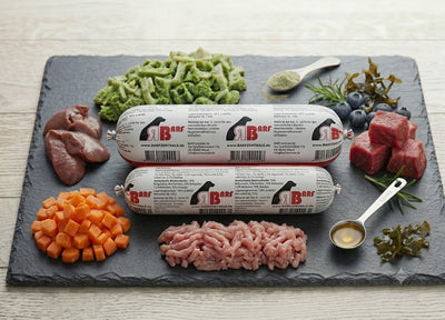 Stimmungsvolles Produktfoto des 'Hausmarke Rindmenü' Hundefutters von BARF-Zentrale. Zwei gestapelte BARF-Rollen (1000g und 500g) liegen auf einer dunklen Schieferplatte, umgeben von frischen, gewürfelten Karotten, rohem Muskelfleisch, grünem Pansen und einem Löffel Lachsöl."